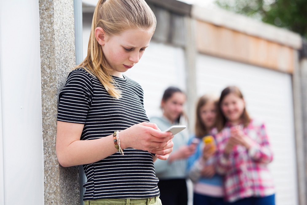 Young girl looking at her phone sadly, while three other girls in the background are staring at her