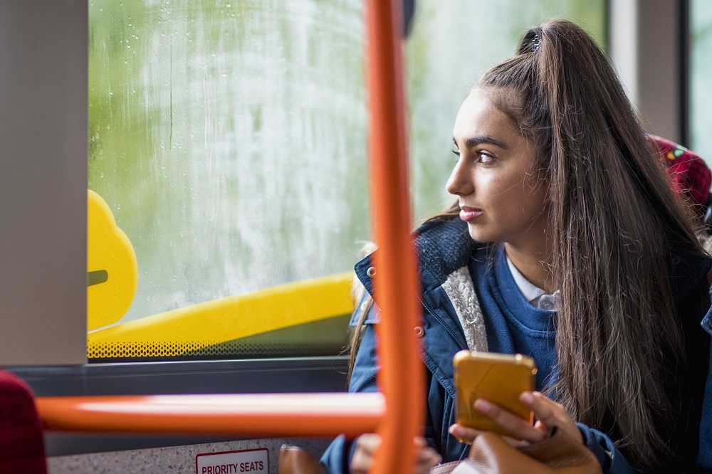 Teen girl sitting on a school bus holding her phone, looking out of the window