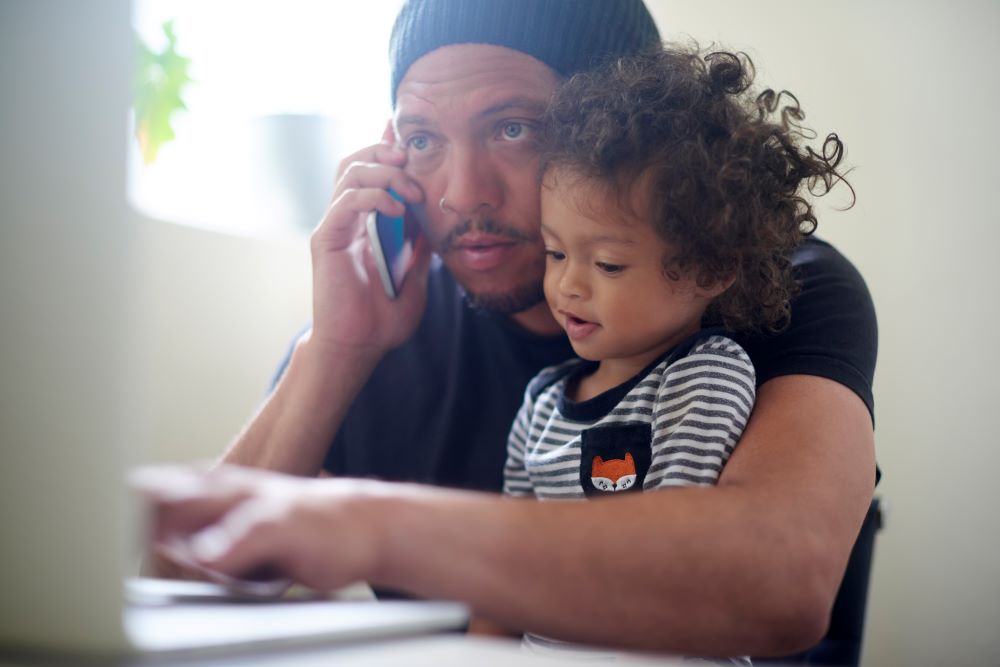 Dad with toddler on his knee, on the phone while looking at a laptop