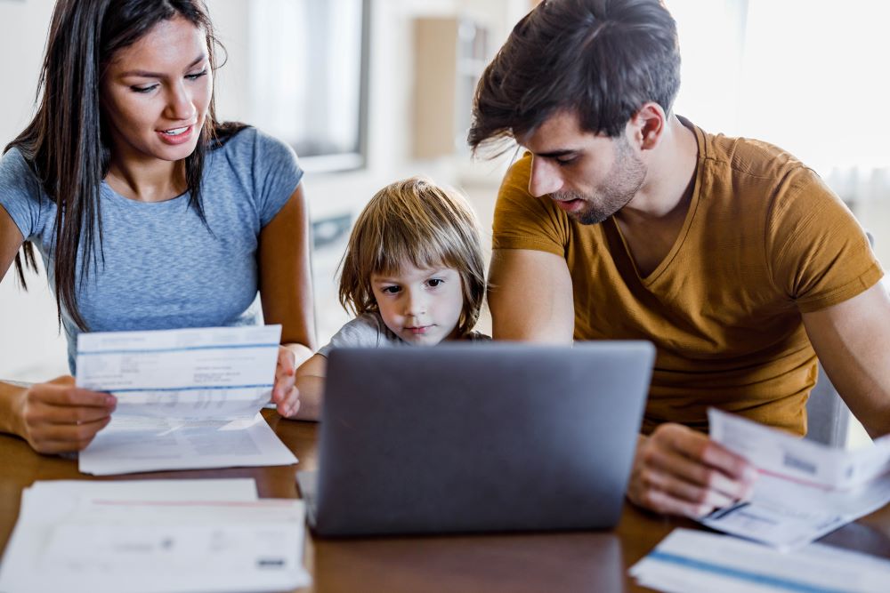 Mum and dad with young child sitting at a table looking at bills with a laptop open in front of them