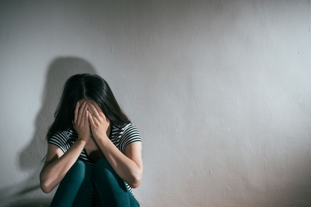 Sad teenage girl sitting on the floor with her back to the wall, covering her face with her hands.