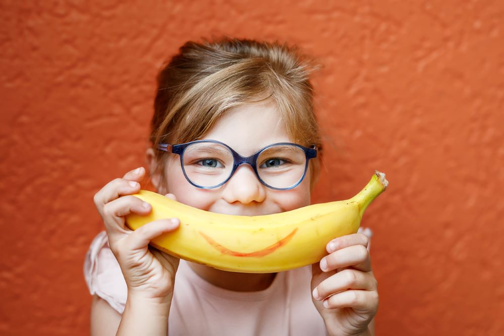 Young girl wearing glasses, holding a banana up to her face like a smile.