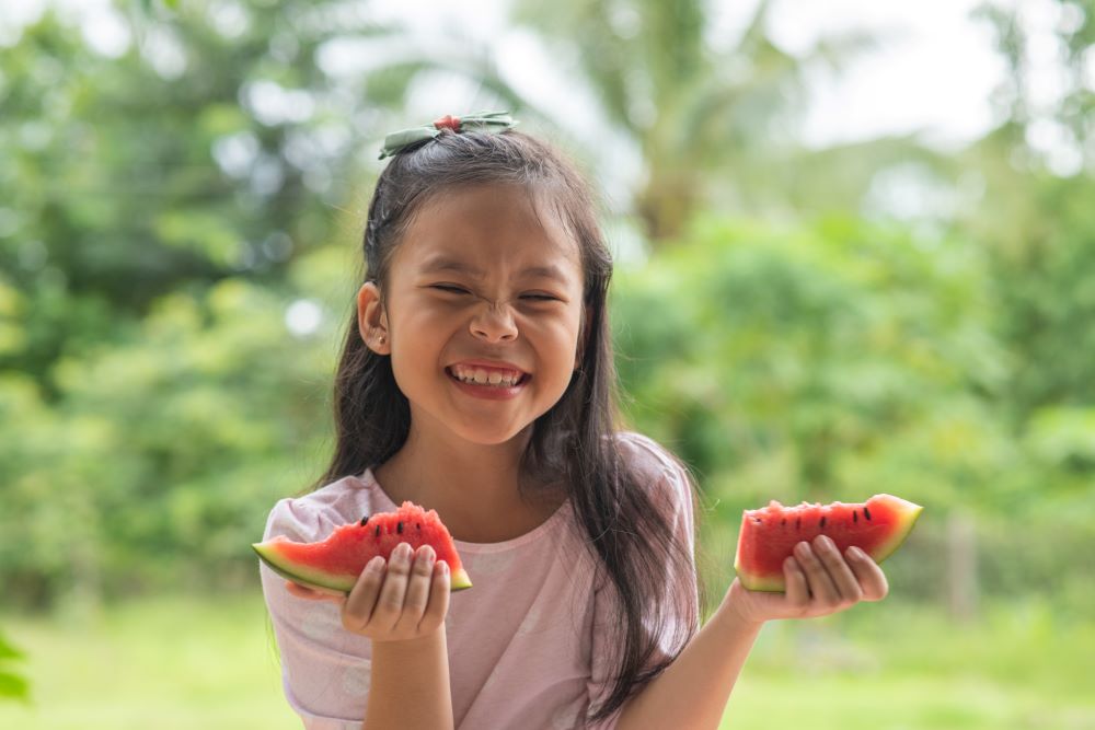 Young girl outdoors, smiling and holding slices of water melon in each hand.