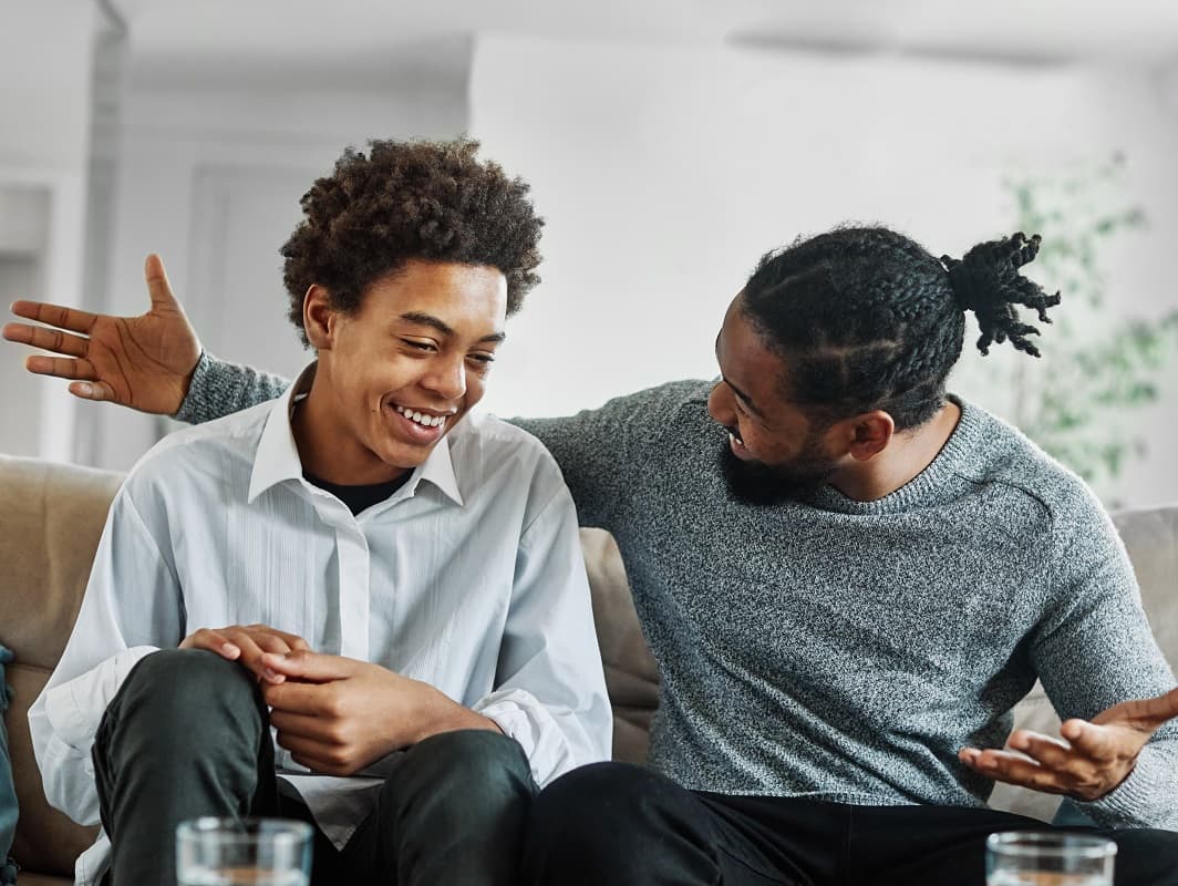 Teen boy and dad sitting on a sofa talking and laughing together.