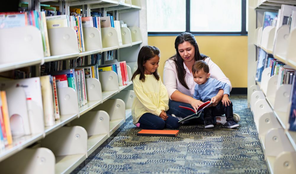 Mother with young child and toddler sitting on the floor reading together in a library.
