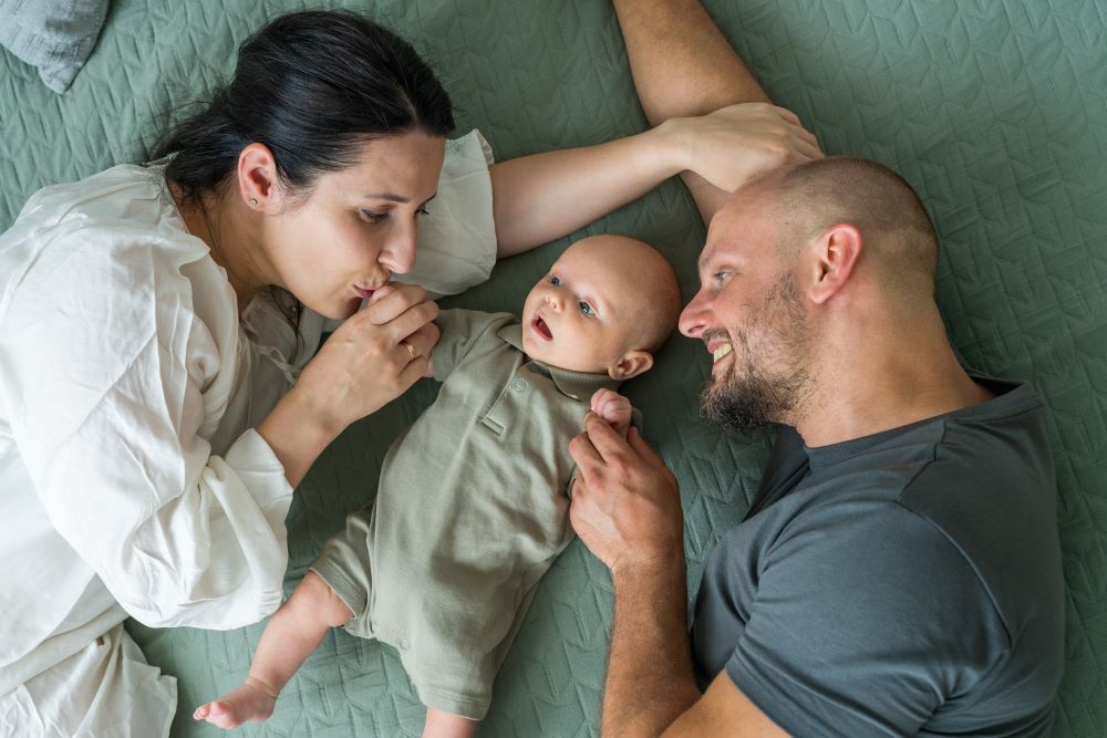 Mum and dad lying on a bed with their newborn baby, looking at the baby lovingly.