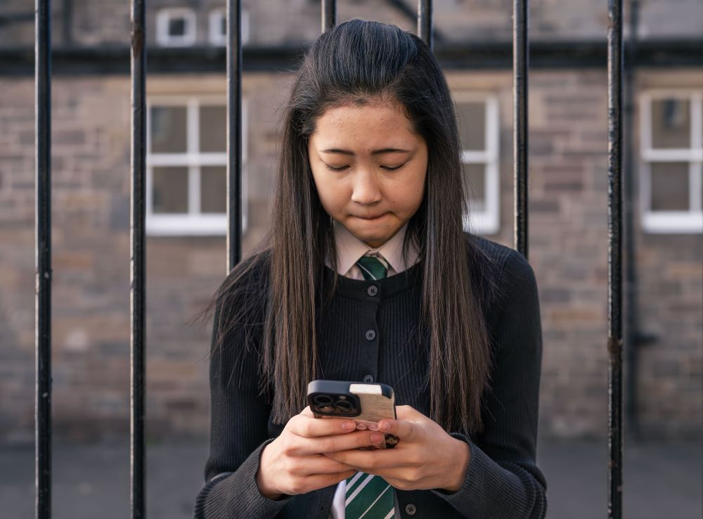 Girl standing outside the school gates looking at her phone and crying.