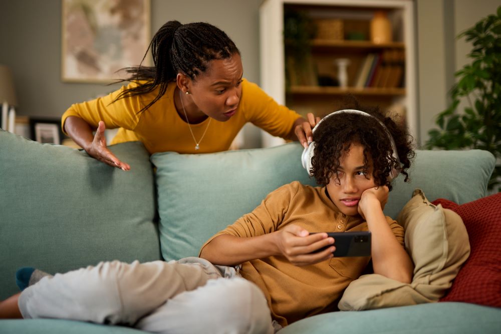 Older child lying on a sofa, looking at their phone with headphones on, while their mum tries to get their attention.