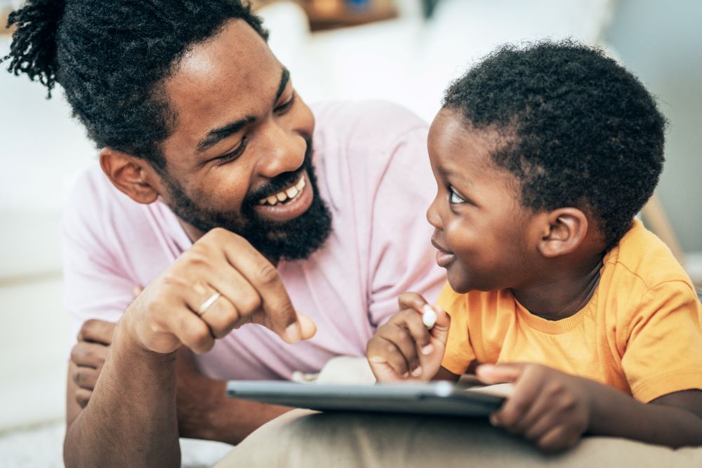 Dad chatting and smiling to his toddler while looking at a tablet together.