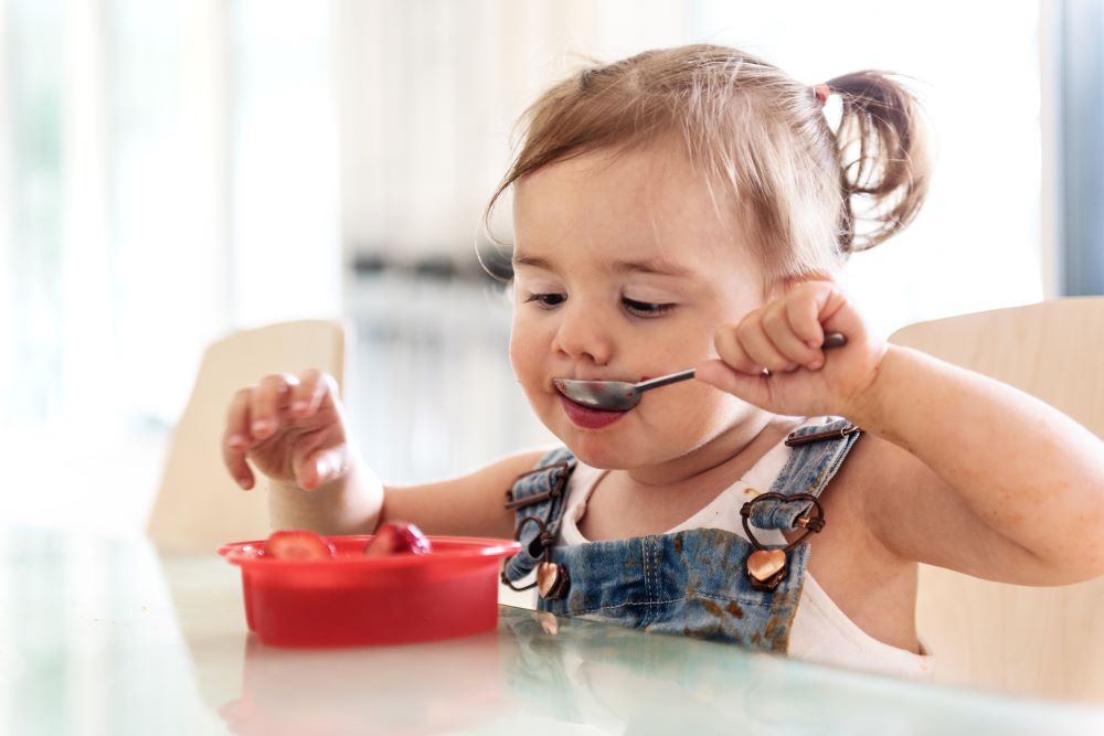 Toddler eating fruit from a bowl with a spoon.