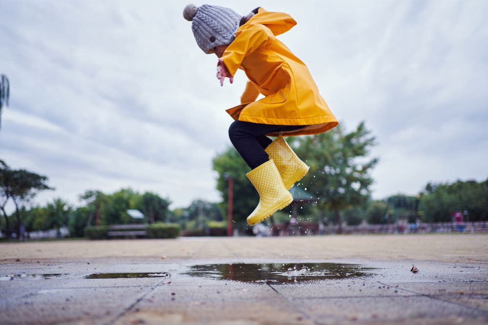 Young child wearing a yellow mac and wellies jumping in a puddle.