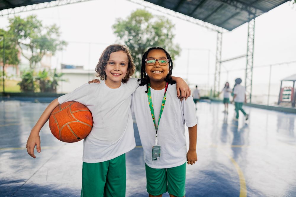 Two girls with their arms around each other, one carrying a netball, the other wearing a sunflower lanyard.