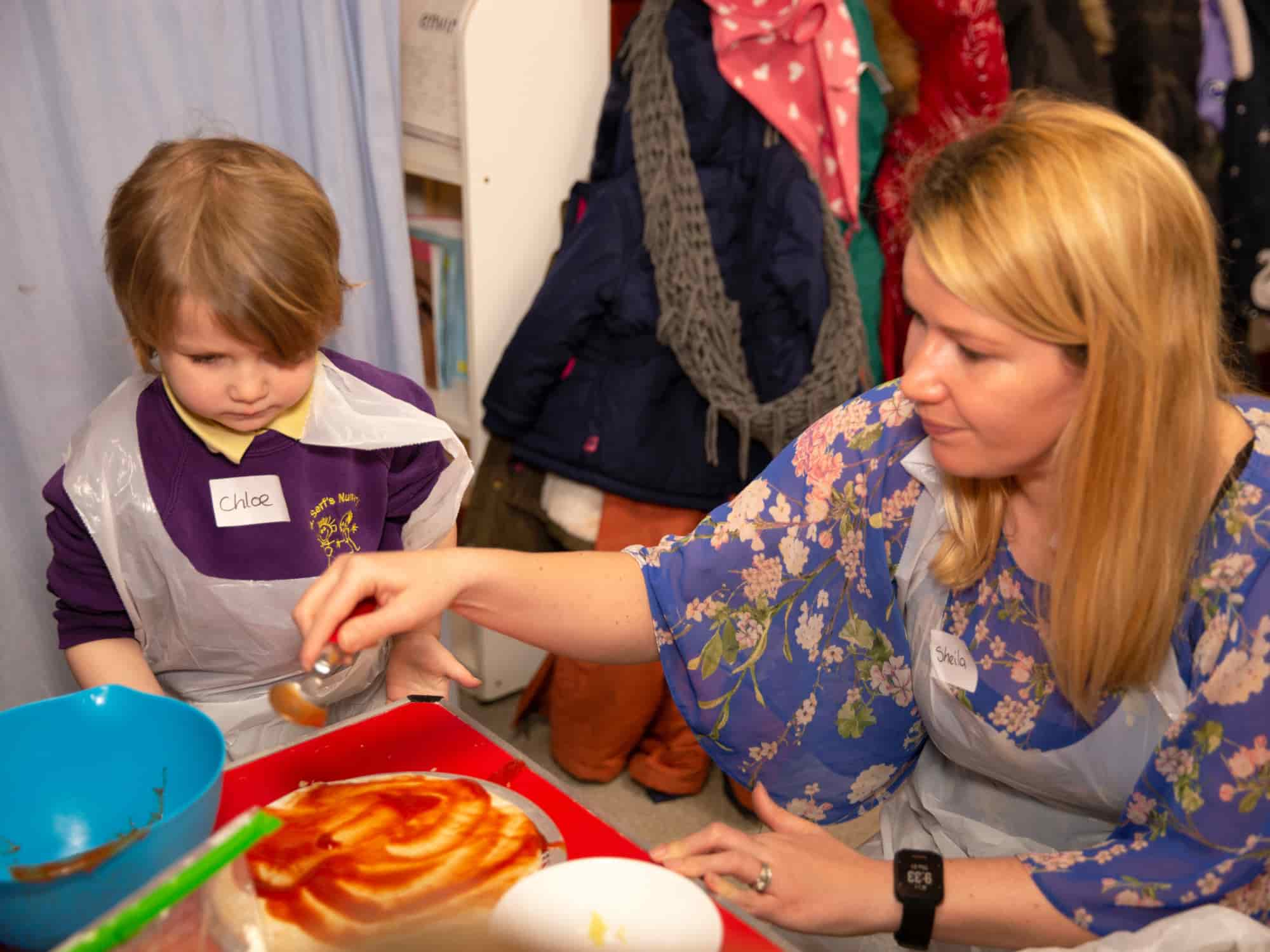 Image of an adult and a child sitting at a table in a playroom, spreading tomato sauce on pizza dough.