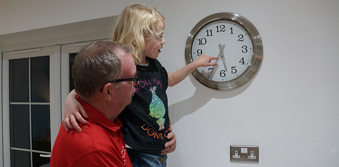 Image of an adult holding a child who is pointing at an analogue clock on the wall.