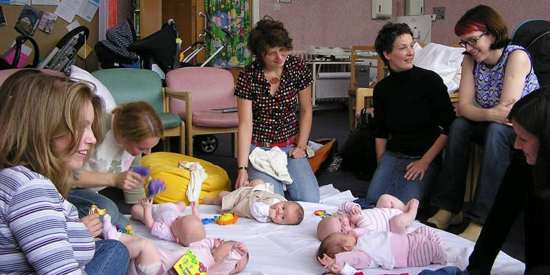 Image of a group of adults sitting in a circle around a blanket with babies lying on it.