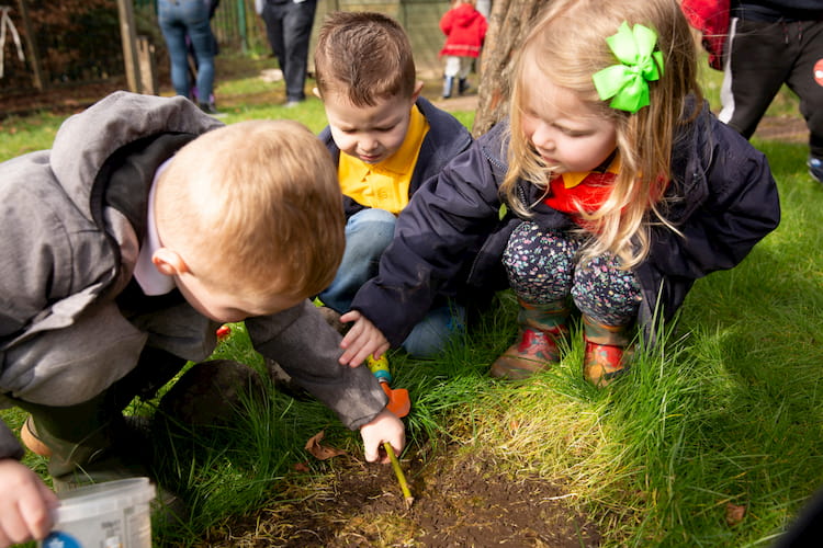 Image of three children crouching on grass, with one child holding a twig.