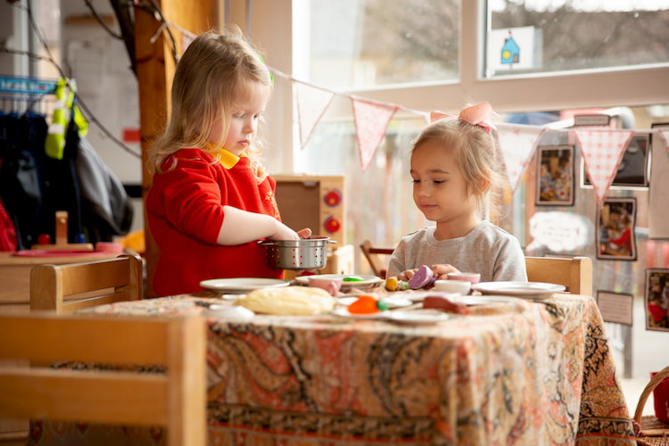 Image of one child sitting at a table while other child serves her toy food.