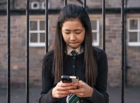 Girl standing outside the school gates looking at her phone and crying.
