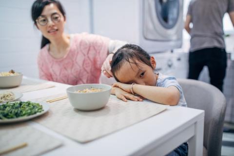 Mother and child sitting at a dinner table, the child with her head in her arms refusing to eat.