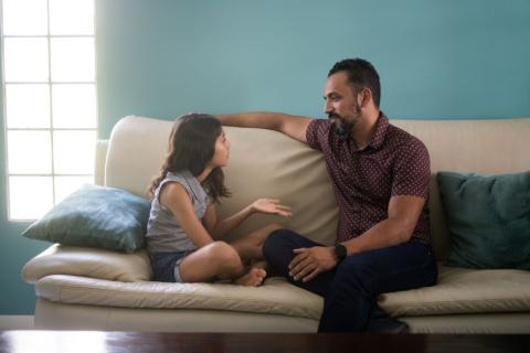 Young girl and dad sitting on a sofa talking together.