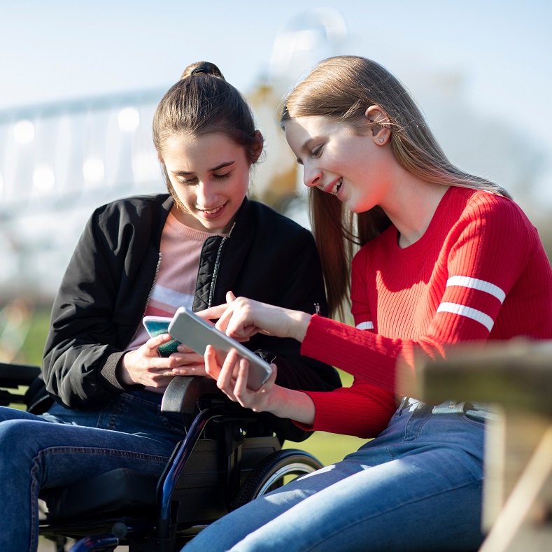 Two teen girls outdoors, looking at their phones. One is in a wheelchair.