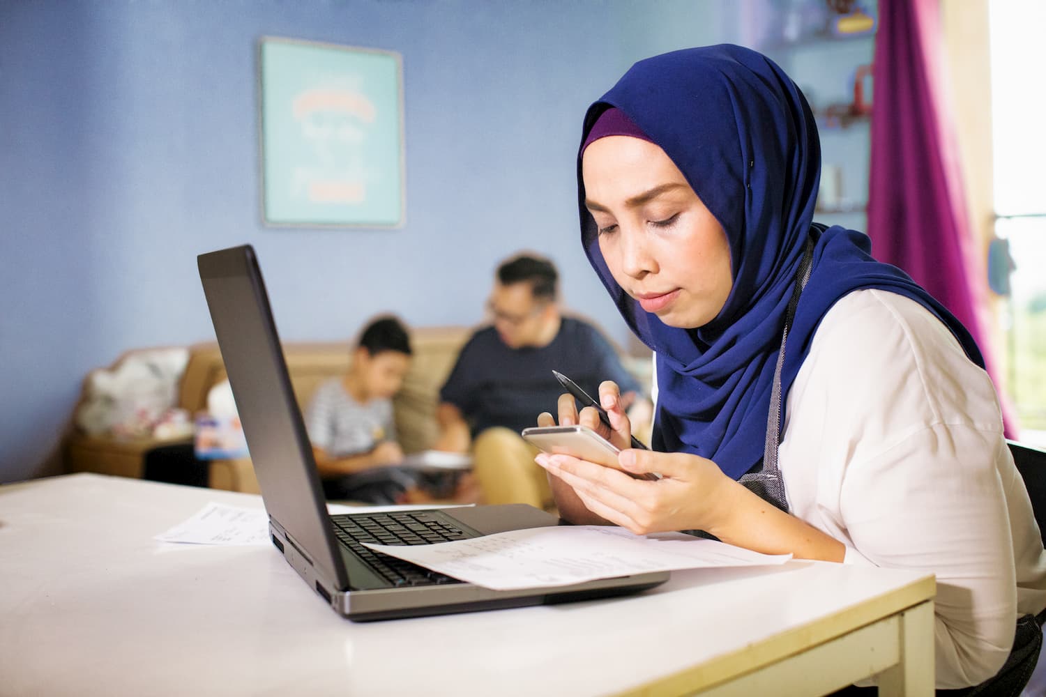 Image of a woman sitting at a table holding a pen and looking at her phone, with a laptop in front of her.